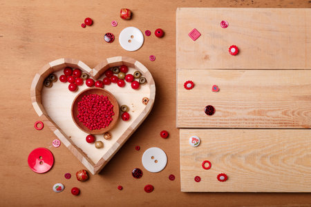 Close up of wooden heart shaped box of red beads and buttons for sewing and embroidery. Set of materials for handcraft, making of bijouterie and accessories. Wooden brown background, copy space.の写真素材