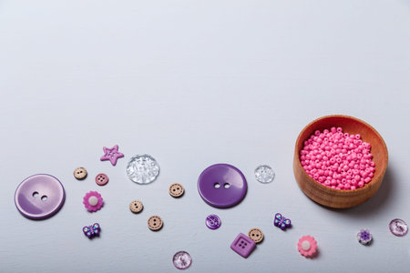 Wooden bowl of pink beads and purple multishaped buttons for sewing and embroidery on white...