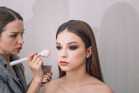 Make-up artist doing visage for beautiful teenage girl, using foundation for face and blush for cheeks. Make up, cosmetics, beauty standards, skin care concept. Close up shot.の写真素材