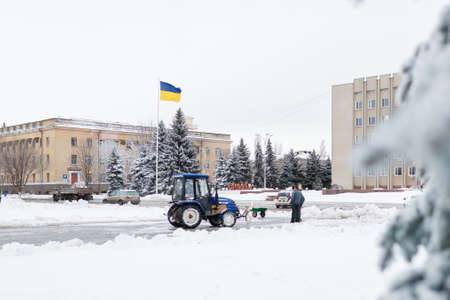 Izmail, Ukraine - December 2019. Removal of snow on town street, working people and tractor. Yellow and blue on building background. Beautiful winter landscape, fir trees covered with snow.のeditorial素材