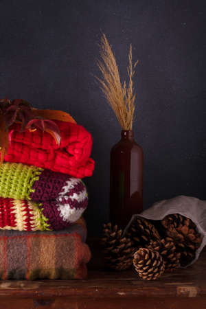 Bright knitted plaids, sweaters, brown vase with dry leaves and pine cones on deep gray background. Autumn composition, hygge conceptの写真素材