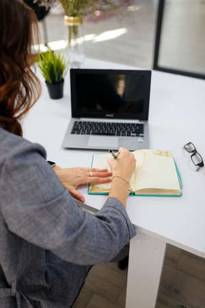Izmail, Ukraine. May 2021. Business woman working in office, writing in notebook back to camera. Laptop on table. Vertical closeup, back viewのeditorial素材