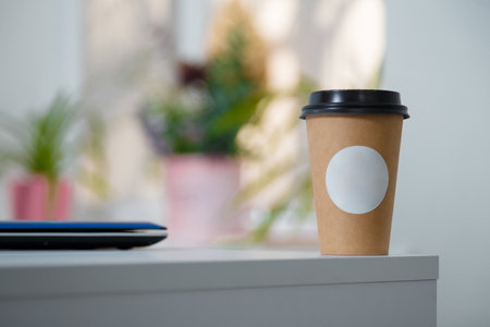 Disposable paper cup of coffee with white mock up label standing on white table. Coffee break at working place. Soft focused close up shot.の写真素材