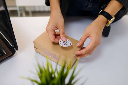 Izmail, Ukraine, May 2021. Female hands sticking a logo label on carton, paper box. Potflower on white table background.のeditorial素材