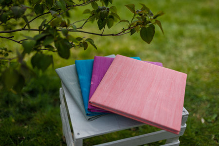 Books, albums, notebooks or photobooks with gray, turquoise, purple and pink leather covers lying on white wooden box among green grass. Soft focused shot.の写真素材