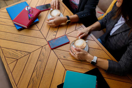 Two colleagues having business lunch, cofee break. Hands holding cappuccino cups. Notebooks, smartphones, pens and glasses on wooden tableの写真素材