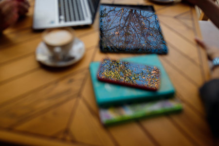 Blue sky and trees reflection in smartphone and tablet on unfocused wooden cafe table. Unfocused...