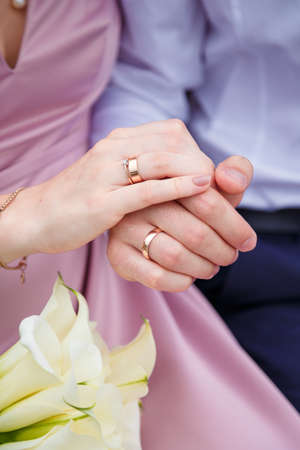 Groom and bride in pink dress holding hands with rings, white calla lilies bouquet. Tender minimalistic style wedding, happy just married. Close up vertical shot.の写真素材