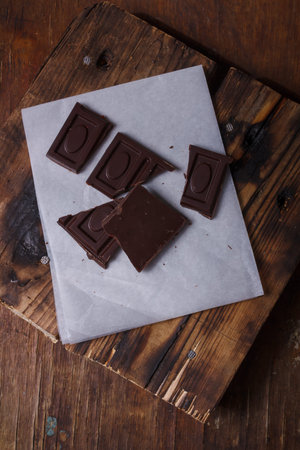 Broken bar of dark bitter chocolate with chilly pepper and red pea pepper inside on white paper, wooden board background. Unusual sweets tasteの写真素材