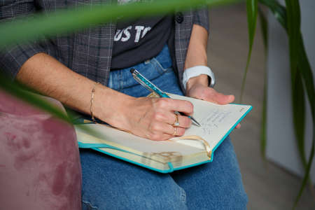 Soft focused close up shot of woman in jeans, gray jacket sitting with notebook, only hands writing with pen visible.の写真素材