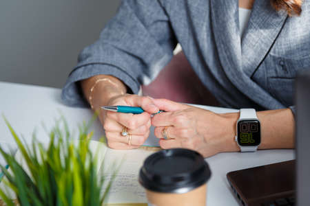 Izmail, Ukraine. May 2021. Business woman in gray jacket holds pen. Hands with watch on table. Laptop, potflower, coffee cup on foreground. Office workのeditorial素材