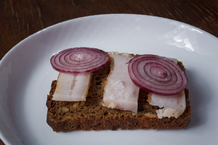 whole grain rye bread and prickled pork lard sandwich with red onions on white plate, wooden table....