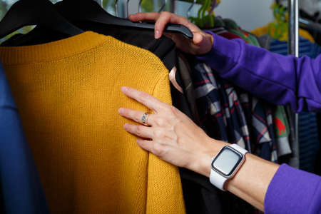 Female hands touching choosing yellow sweater in shop. Racks with clothes on background. Soft focused closeup shotの写真素材