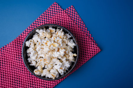 Heap of popcorn in black bowl on bright pink mat, blue background. Top view, copy space.の写真素材