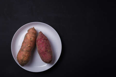 Orange, purple raw sweet potatoes, yam on white plate, black table. Farm harvest, organic vegetables, shop local vegan food. Top view, copy spaceの写真素材