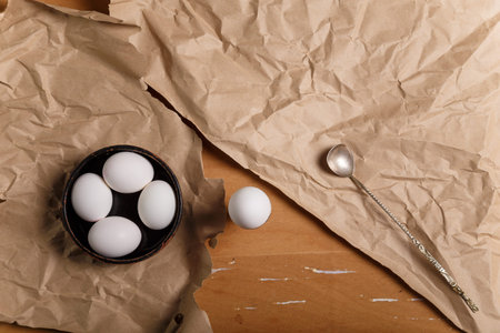 Boiled eggs in black bowl and in egg cup with silver spoon on paper on wooden table. Lunch or Easter...