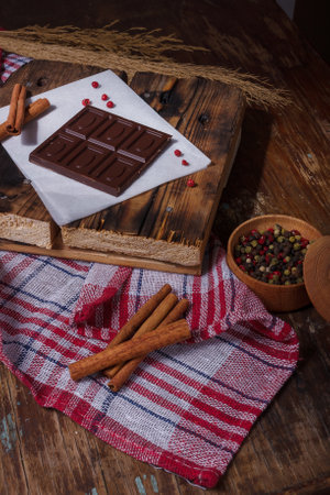 Broken bar of dark bitter chocolate with chilly pepper, cinnamon, red pea pepper inside on paper, wooden board background. Unusual tasteの写真素材