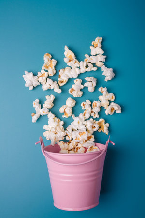 Small pink bucket on blue background with popcorn spilled out, vertical shot