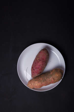 Orange, purple raw sweet potatoes, yam on white plate, black table. Farm harvest, organic vegetables, shop local vegan food. Top view, copy spaceの写真素材