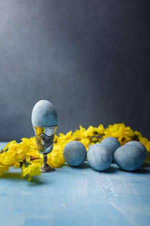 Blue painted egg in silver egg cup, easter eggs and yellow flowers on gray background. Ukrainian Easter. Vertical shot, copy spaceの写真素材