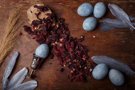 Beautiful diy blue painted easter eggs, egg cup, dry Hibiscus tea and silver decorative feathers on wooden background, top viewの写真素材