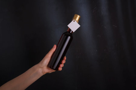 Hand holding bottle of red wine with template label, no brand. Alcohol beverage on black background....
