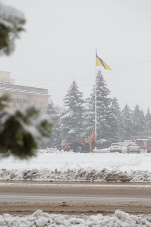 Izmail, Ukraine - February 2022. Removal of snow on town street, working tractor. Yellow and blue flag, building on background. Beautiful winter landscape, fir trees covered with snow.のeditorial素材