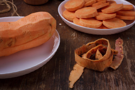 Orange raw sweet potato, skin, yam slices on plate, wooden rustic table. Farm harvest, organic vegetables, shop local, diet, nutritionの写真素材