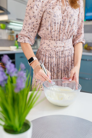 Woman whipping egg whites with manual whisk in glass bowl, making cream, mousse or meringue for dessert. Flowers on foregroundの写真素材