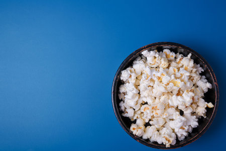 Heap of popcorn in black bowl on bright blue background. Top view, copy space.の写真素材