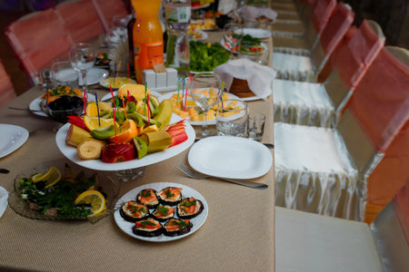 Festive table with appetizers. Fruit plate with banana, kiwi and citrus. Herring with lemon and greenery. Eggplants with sauce and tomatoes. Empty glasses, plates for guestsの写真素材