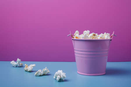 Small pink bucket of pop corn on blue and pink background. Some popcorns lying near. Copy space.の写真素材