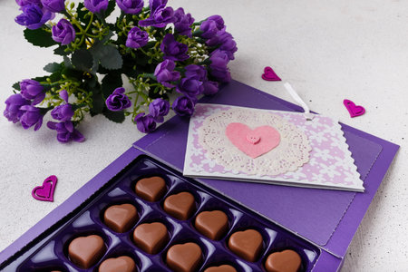 Violet box of heart shaped chocolates, postcard and flowers on white background. Valentines day 14...