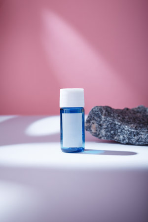 Small blue bottle with blank label, no brand mockup on white table, pink backdrop, decorative stone in harsh light. Care cosmeticsの写真素材