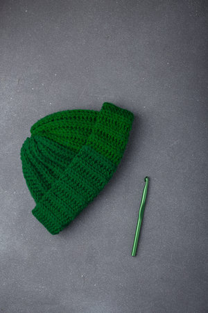 Green crochet beanie cap or hat on gray background. Crocheting hook near. Top view, copy space. Vertical shotの写真素材