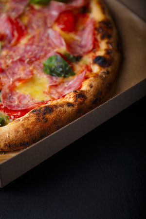 Pizza with cheese, ham, green basil leaves and tomatoes in paper box on black background. Food product advertising banner, copy space.の写真素材