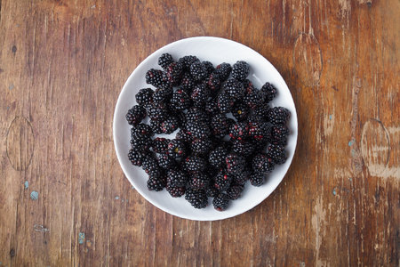 ripe fresh blackberries in white plate on brown old rustic wooden table, top view, copy space.
