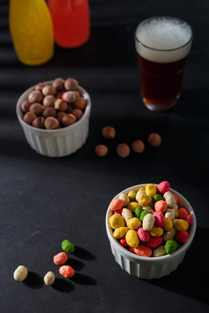Multiflavored peanuts in bright multicolored icing in white bowl on black table background. Beer and non alcohol drinks, hazelnuts. Vertical shot.