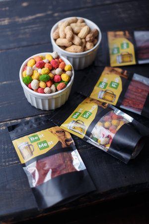 Izmail, Ukraine. October, 2023. Package of multiflavored peanuts in bright multicolored icing on black wooden background. Some unpeeled nuts in bowl.