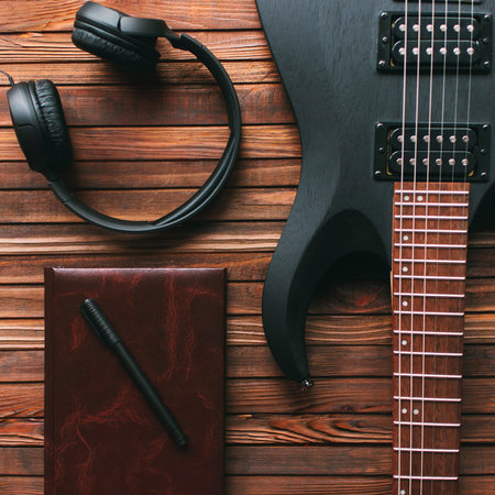 Black electric guitar, notepad and headphones on a wooden table. Making music concept.
