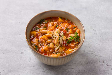 Vegan seasonal italian minestrone soup with pumpkin, beans, tomatoes, bell peppers, pasta and herbs on gray textured background. Delicious homemade foodの写真素材
