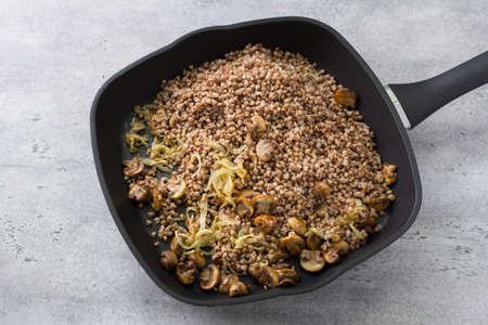 Non-stick frying pan with buckwheat, fried onions and champignon mushrooms on a gray textured concrete background, top view, homemade food. Cooking a traditional Jewish dish kasha varnishesの写真素材