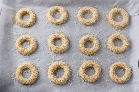 Raw rings of greek orange sesame cookies koulourakya on a parchment baking sheet before baking, top view. Step by step instructions. Do it yourself. Step 7. Place raw cookies on a baking sheet, brush with egg and sprinkle with sesame seedsの写真素材