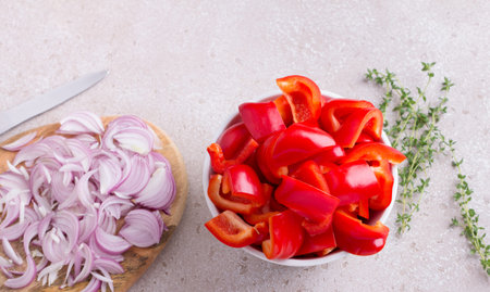 A bowl of chopped red sweet peppers, a wooden board with chopped red onions, fresh thyme sprigs and a kitchen knife on a beige textured background, top viewの写真素材
