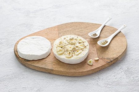 Wooden board with a cut head of white mold cheese, chopped garlic and spices on a light gray background. Stage of cooking a delicious holiday snackの写真素材