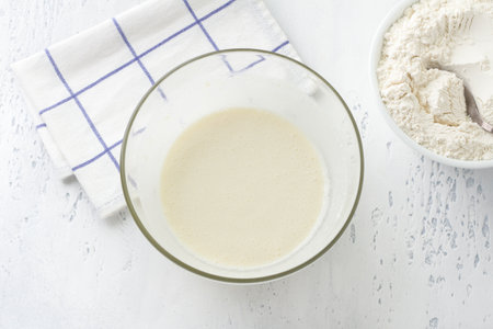 A glass bowl with a whipped mass of eggs, butter, sugar and milk on a light blue background, top view. Cooking homemade milk cakes, do it yourself, step by step, step 5.の写真素材