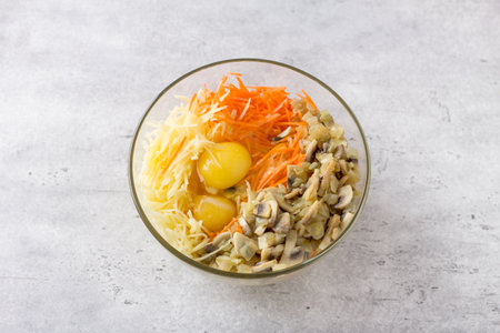 Glass bowl with grated potatoes, carrots, eggs and fried mushrooms with onions for making delicious potato pie, gratin or casserole on gray textured background, top view.の写真素材