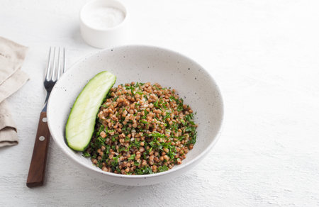 White bowl with boiled buckwheat and kale on a gray background, served with fresh cucumber. Healthy homemade vegan foodの写真素材