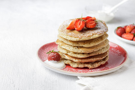 Stack of homemade pancakes with fresh strawberries and powdered sugar on plate, served with yogurt,...