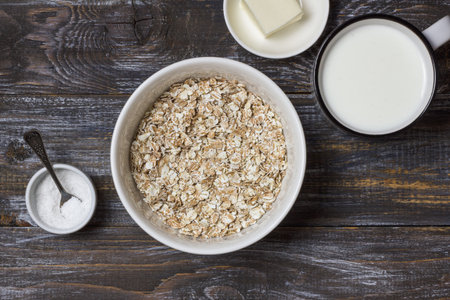 Raw four grain flakes in a ceramic bowl with milk, butter and salt arranged on a rustic wooden table, showing natural ingredients for healthy porridgeの写真素材